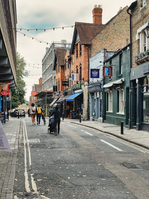 A street scene in a residential and commercial area during daytime, with narrow pavement and road, lined with multicoloured brick and plaster buildings featuring shopfronts and storefront signage. Visible storefronts include a wine shop and other retail outlets, with awnings and window displays. Pedestrians are walking along the pavement, some pushing strollers and carrying shopping bags. Overhead, string lights are suspended across the street, indicating a lively environment. The setting suggests a typical urban high street that is possibly part of a relocation route in Beckenham. The scene does not show any moving or packing activity directly, but the context aligns with house removals and home relocation processes. The lighting is natural, and the street appears quiet with light foot traffic, possibly during mid-morning or early afternoon.