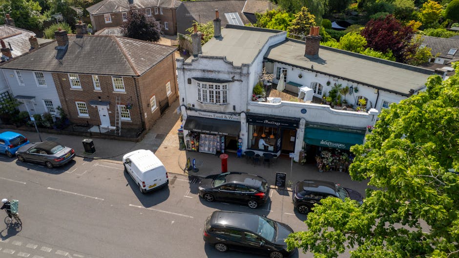 An aerial view of a small high street in Beckenham showing a row of buildings with retail shops, including a charity shop and a florist, situated along the pavement. Parked vehicles, including a white van, a black hatchback, and other cars, are aligned along the curbside in front of the shops. The white van is positioned closest to a door, possibly being loaded or unloaded, indicating a home relocation process. The scene also features green foliage from trees lining the street, with sunlight casting shadows on the pavement and buildings, suggesting it is daytime. This image captures the typical environment involved in house removals and furniture transport within a local area, with visible packaging materials, such as cardboard boxes near the shop entrances, emphasizing packing and moving logistics. The setting underscores the importance of efficient loading and parking considerations during local moves managed by professionals like Man and Van Beckenham.