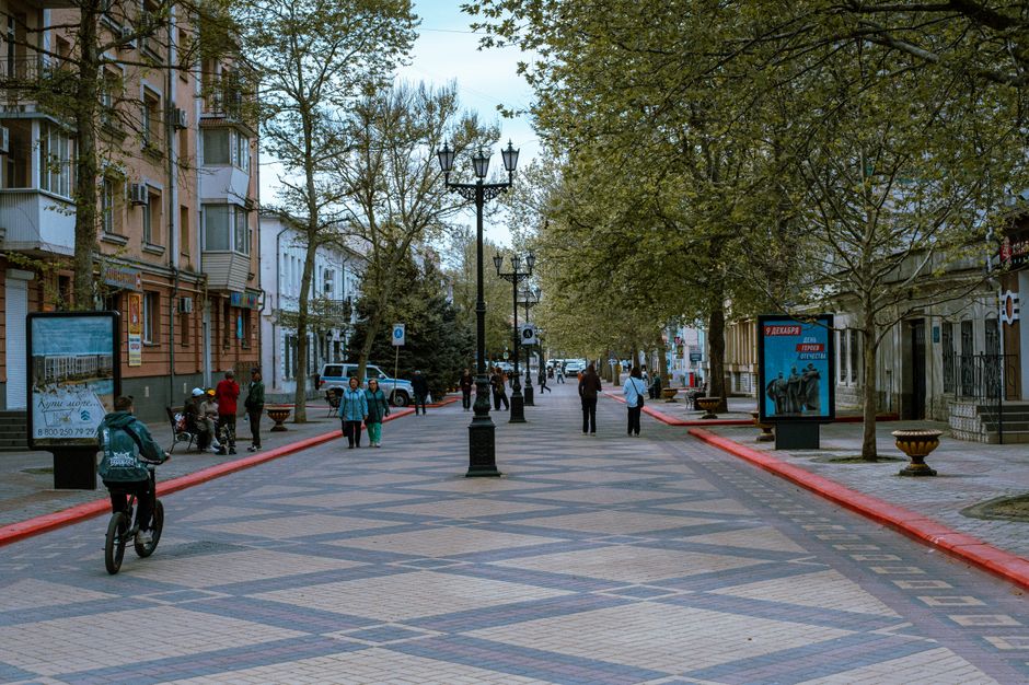 A street scene in a residential and commercial area during daytime, with narrow pavement and road, lined with multicoloured brick and plaster buildings featuring shopfronts and storefront signage. Visible storefronts include a wine shop and other retail outlets, with awnings and window displays. Pedestrians are walking along the pavement, some pushing strollers and carrying shopping bags. Overhead, string lights are suspended across the street, indicating a lively environment. The setting suggests a typical urban high street that is possibly part of a relocation route in Beckenham. The scene does not show any moving or packing activity directly, but the context aligns with house removals and home relocation processes. The lighting is natural, and the street appears quiet with light foot traffic, possibly during mid-morning or early afternoon.