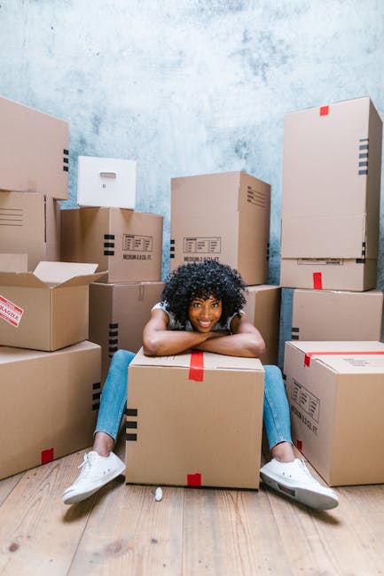 In a room with a textured light blue wall and wooden flooring, a woman with curly black hair is sitting on the floor, smiling and leaning on a medium-sized cardboard box secured with red packing tape. Surrounding her are various stacked and scattered cardboard boxes, some sealed with tape and others partially open, revealing packing materials. The boxes vary in size and are arranged both on the floor and against the wall, indicating a home relocation or packing process. The scene is well-lit with natural or soft indoor lighting, and the woman is dressed casually in a white top and blue jeans with white sneakers, reflecting a moving or packing operation. This setup aligns with the activity of furniture transport, packing and moving, and loading processes handled by Man and Van Beckenham, supporting professional removals services.