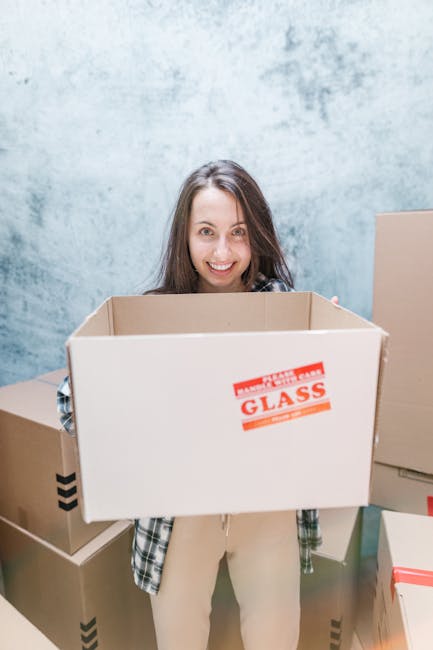 A woman smiling while holding an open cardboard box labeled 'GLASS' inside a room with a painted blue wall. The box contains some packing materials and is part of a home relocation or moving process. Surrounding her are additional closed cardboard boxes of various sizes, some with black tape or red tape, which are positioned on the floor and against the wall. The scene suggests packing or unpacking activities related to furniture transport or house removals, consistent with the services offered by Man and Van Beckenham. The lighting is bright and even, highlighting the packaging and the person's cheerful expression during the loading or moving preparations.