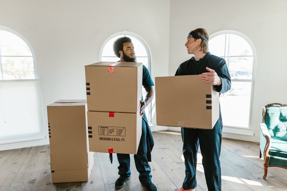 In a room with a textured light blue wall and wooden flooring, a woman with curly black hair is sitting on the floor, smiling and leaning on a medium-sized cardboard box secured with red packing tape. Surrounding her are various stacked and scattered cardboard boxes, some sealed with tape and others partially open, revealing packing materials. The boxes vary in size and are arranged both on the floor and against the wall, indicating a home relocation or packing process. The scene is well-lit with natural or soft indoor lighting, and the woman is dressed casually in a white top and blue jeans with white sneakers, reflecting a moving or packing operation. This setup aligns with the activity of furniture transport, packing and moving, and loading processes handled by Man and Van Beckenham, supporting professional removals services.