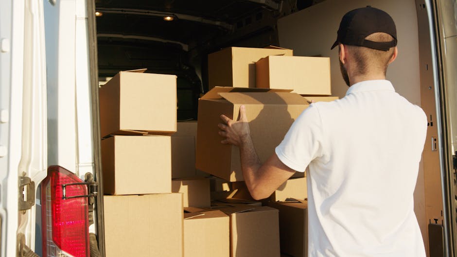 A man wearing a dark jacket and light-colored cap is loading cardboard boxes into the open rear of a white moving van parked beside a modern building with large glass windows and a grey exterior. The
